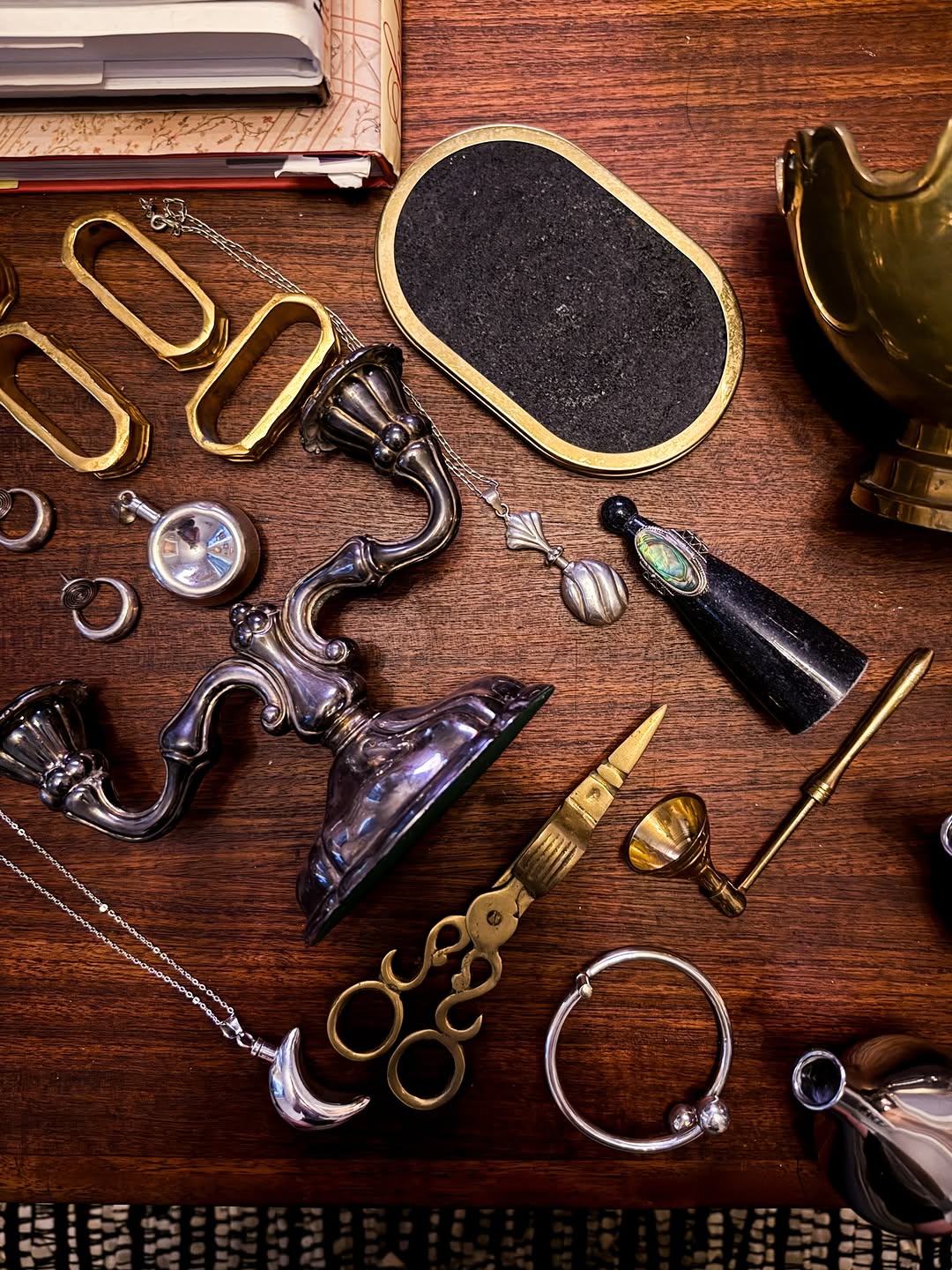 A styled flat-lay of vintage and antique jewelry and accessories arranged on a dark wooden table, including silver and gold rings, necklaces, a candlestick, a small mirror, and other decorative pieces.