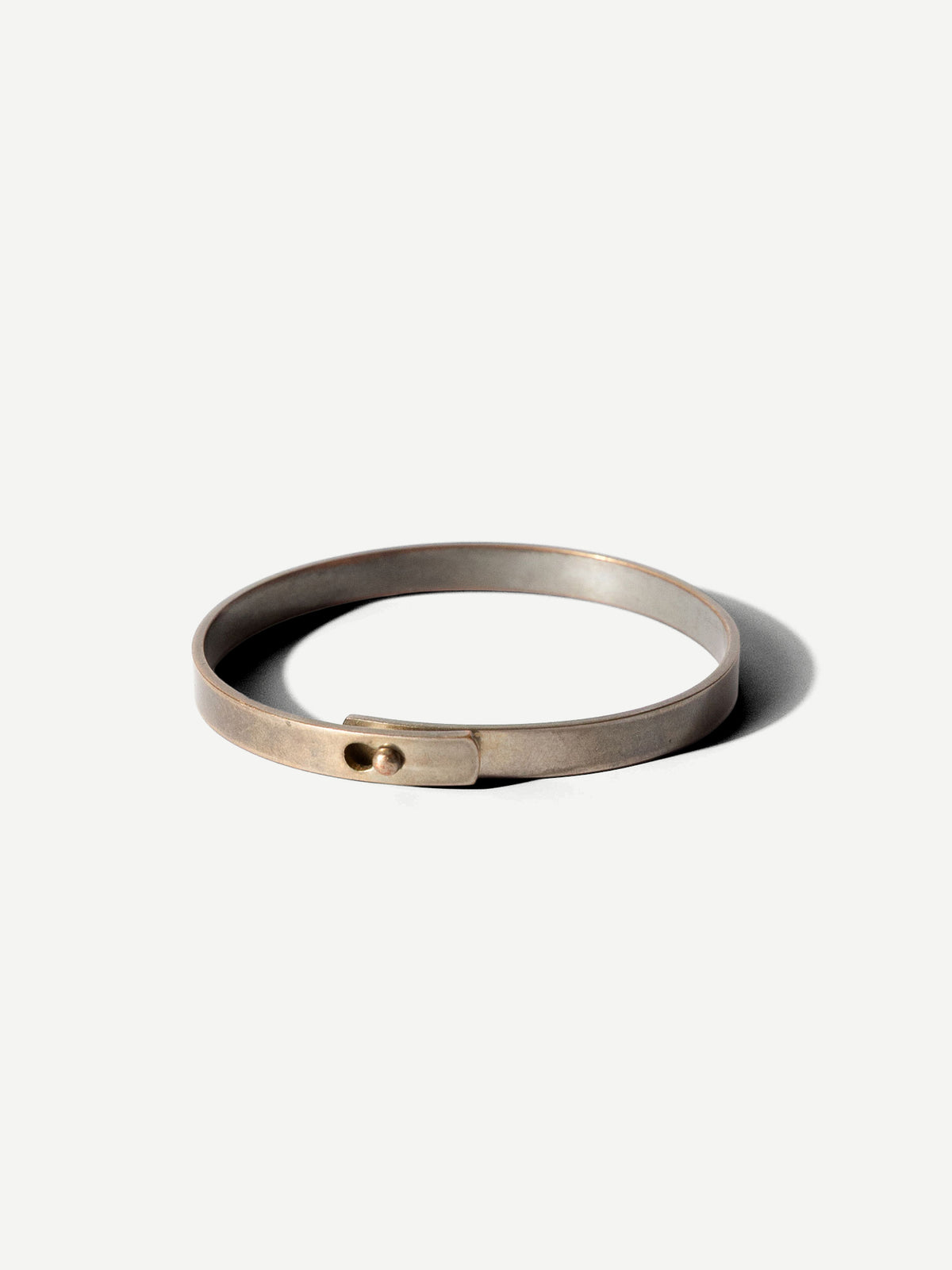 A minimalist, matte metallic cuff bracelet with a small circular detail on the front, displayed on a clean off-white background with a soft shadow.